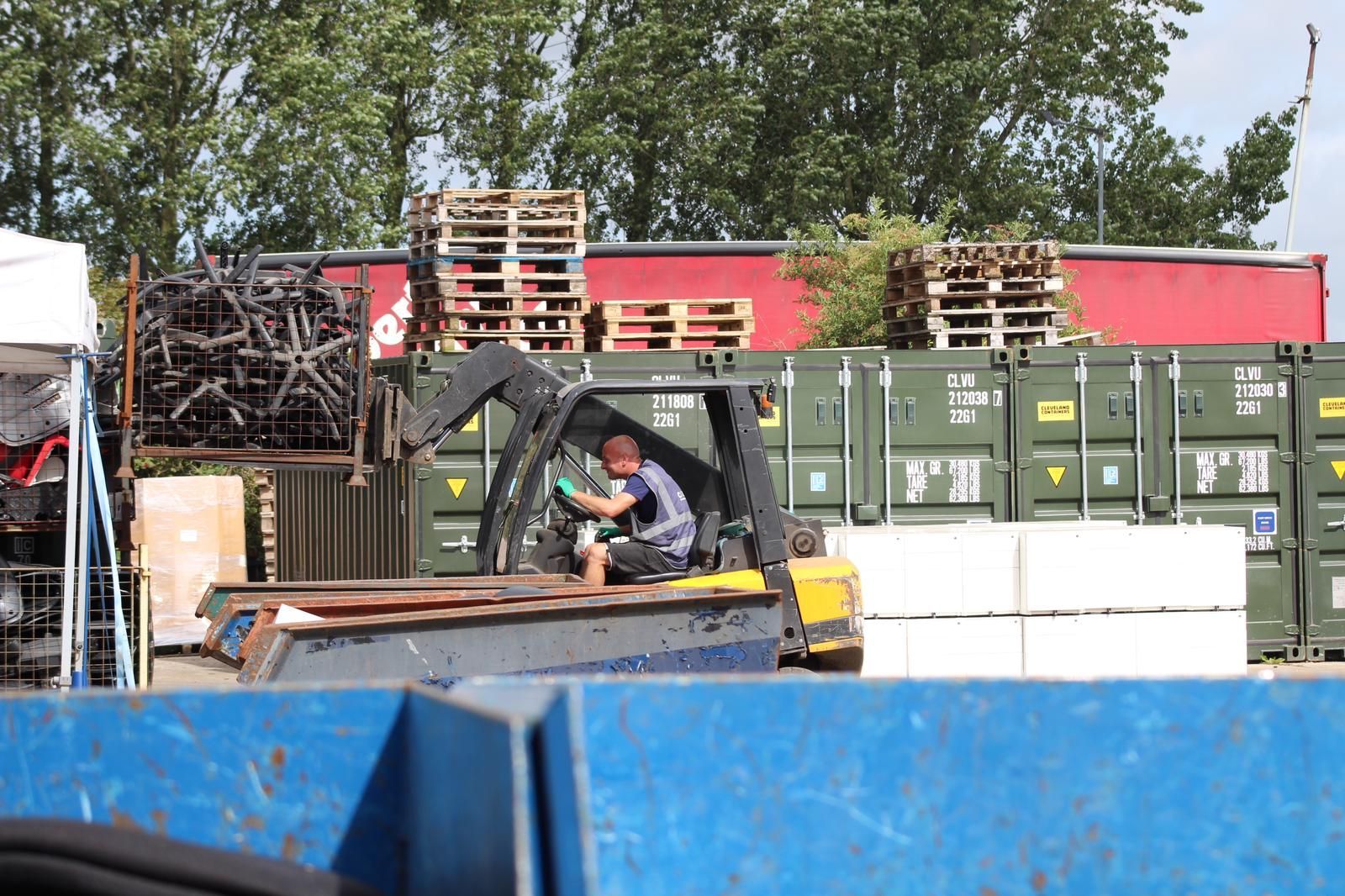 A person operating a forklift in a storage yard, moving items near containers and pallets.