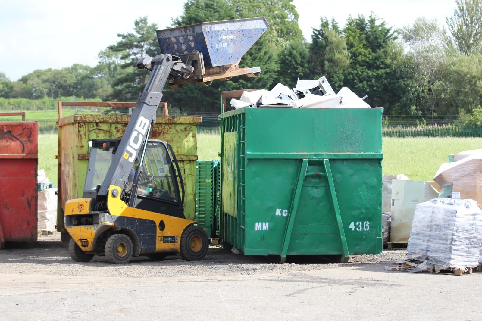 Yellow JCB forklift dumping white debris into a large green dumpster at a recycling facility.