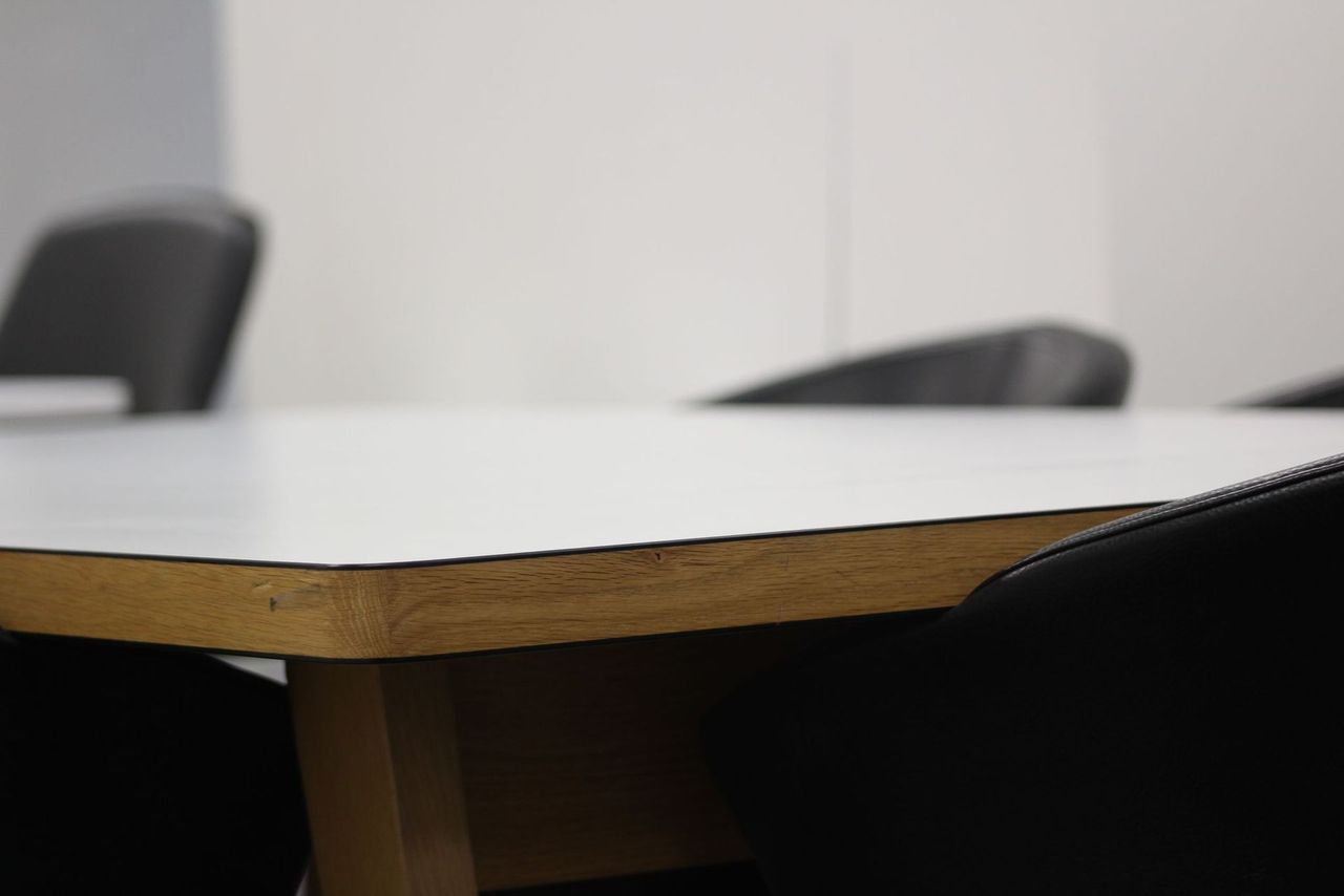 Close-up of a white table edge with brown trim, surrounded by black chairs in a room.