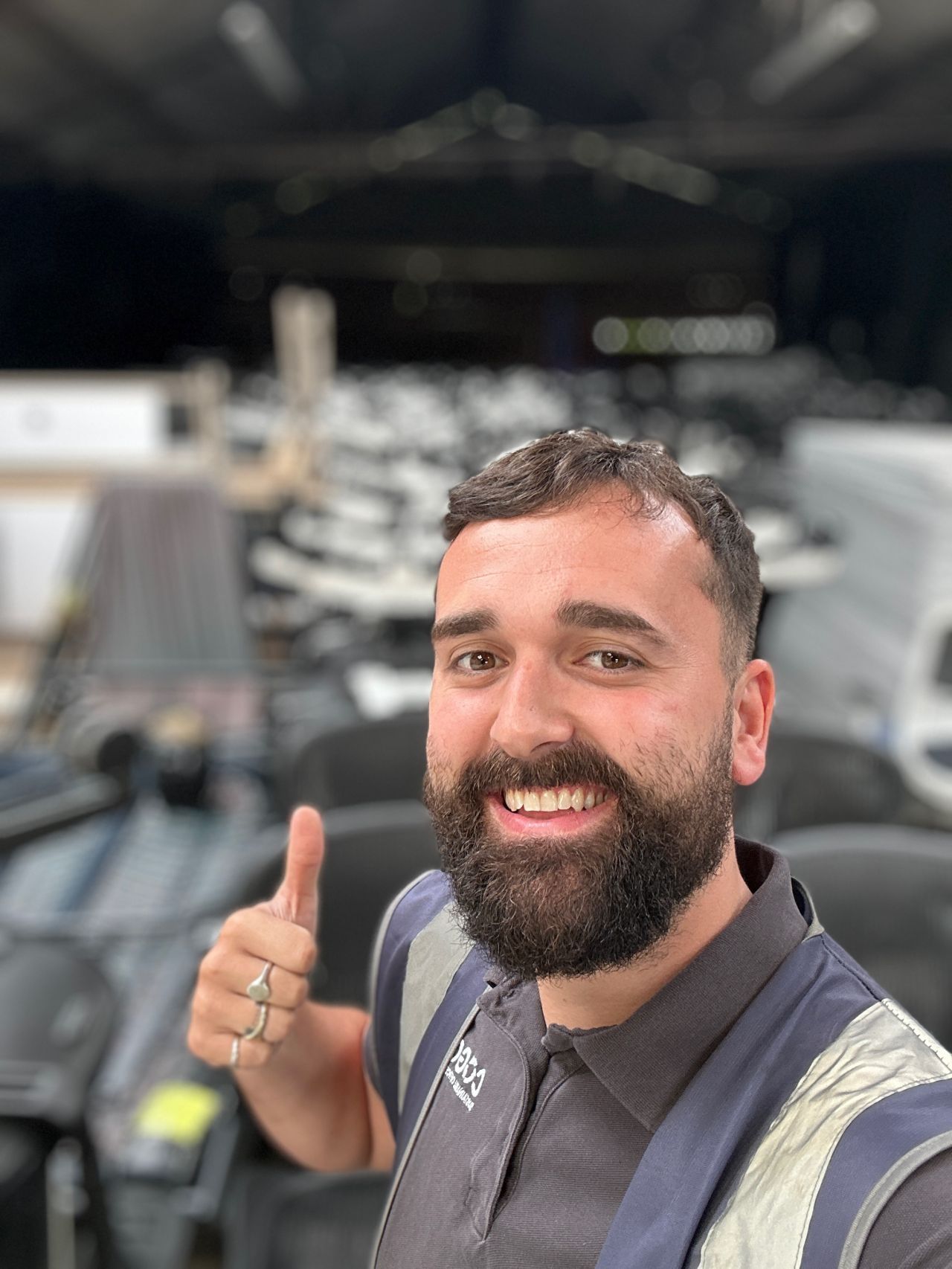Man with a beard giving a thumbs up, smiling. Wearing a work vest, with a blurred warehouse background.