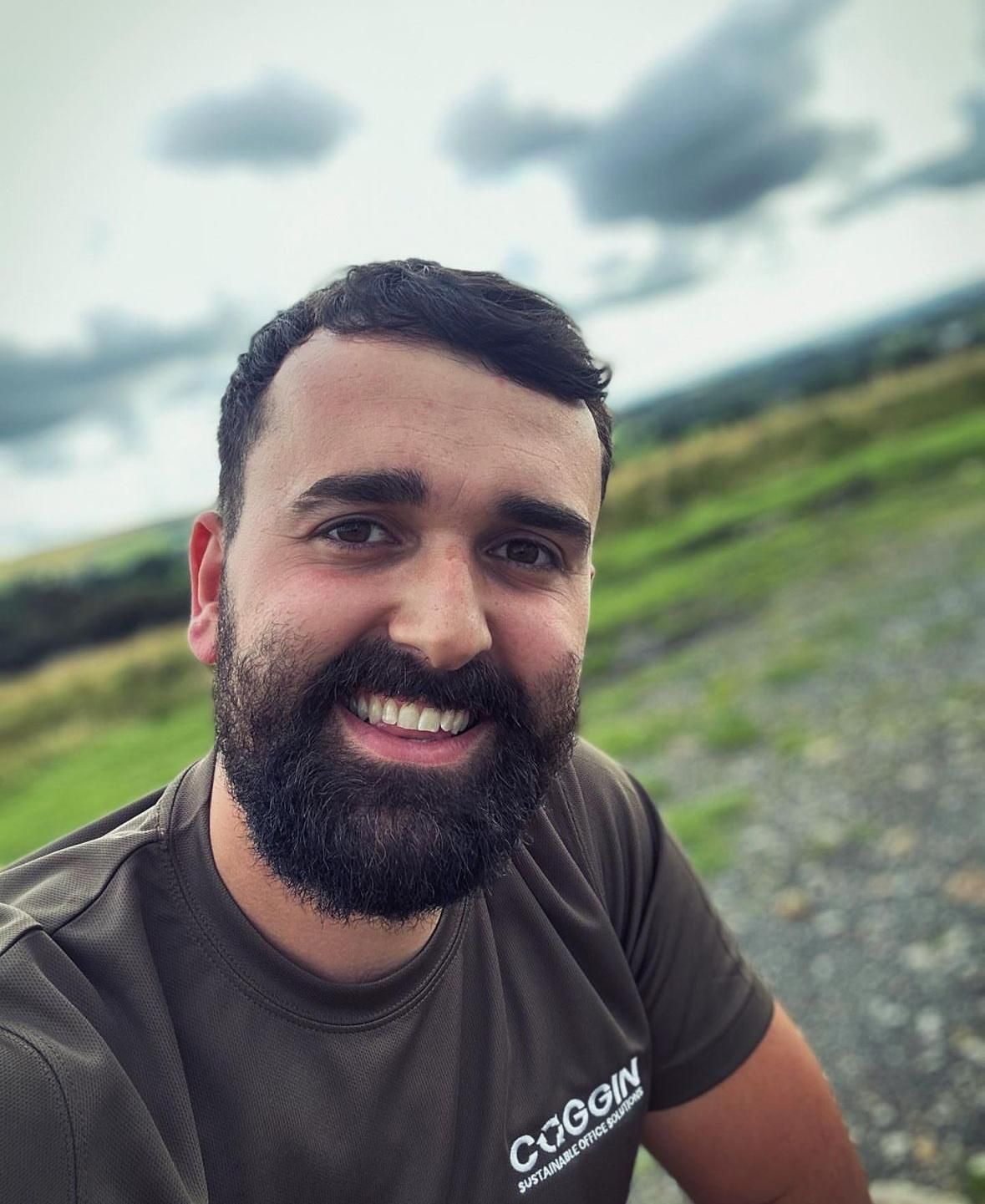 Man with a beard smiling outdoors on a hillside under a cloudy sky.