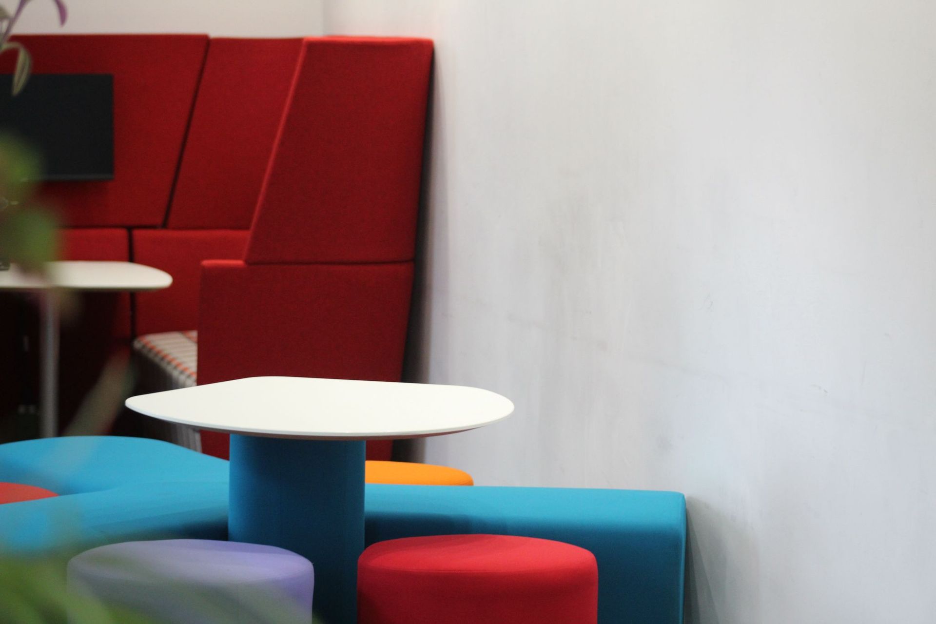 A modern office lounge area with red, blue, and white seating and a white table against a white wall.