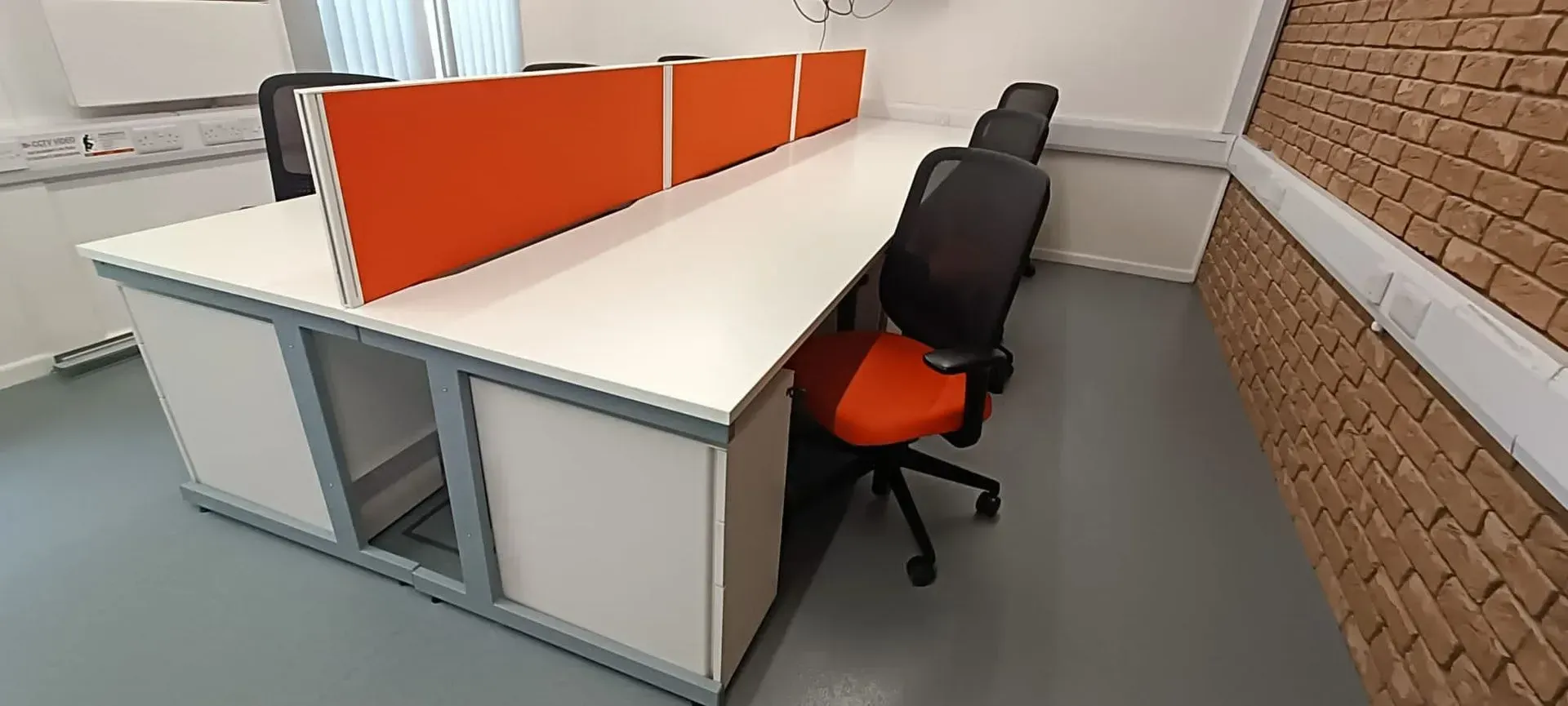 Office with shared desks, orange dividers, and rolling chairs. Brick wall and gray floor visible.