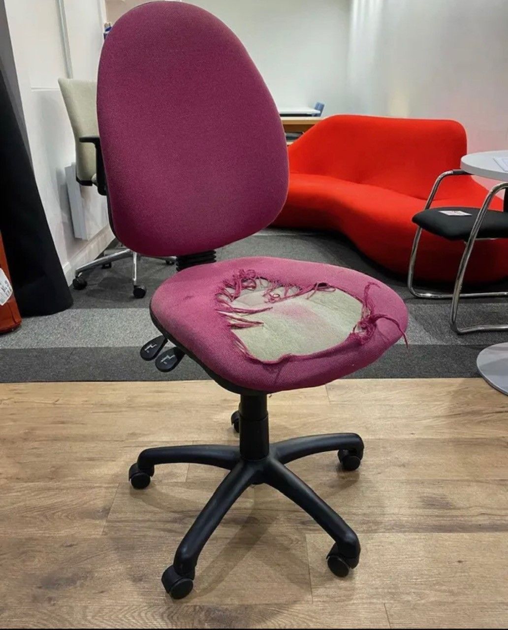 Damaged magenta office chair with torn seat, sitting on a wooden floor in an office.