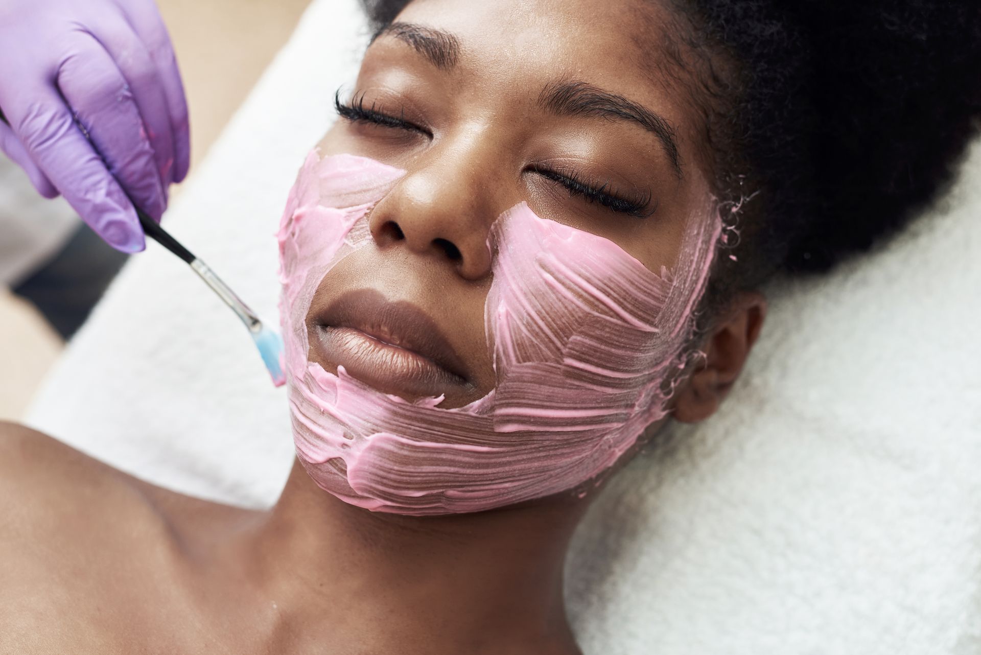 A woman is getting a pink mask on her face.
