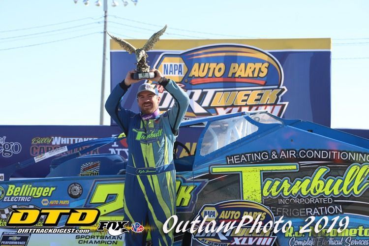 Race car driver holding trophy; car in the background; outdoor setting.