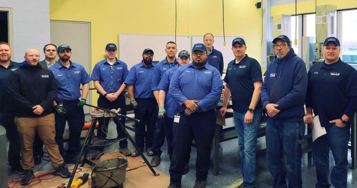 Group of men in blue shirts and hats in a workshop setting, posing for a photo.