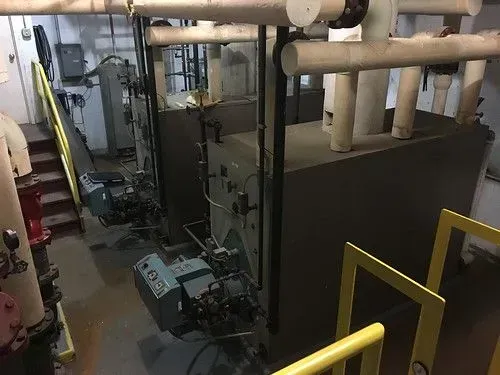 Industrial boiler room with large black HVAC units, pipes overhead, and yellow safety rails on a platform