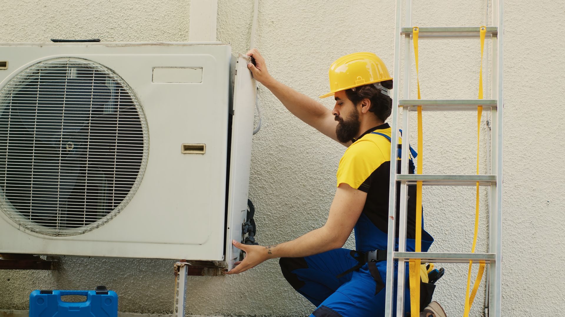A technician in a yellow hardhat servicing an outdoor HVAC condenser unit beside a ladder. A technician in a yellow hardhat servicing an outdoor HVAC condenser unit beside a ladder.
