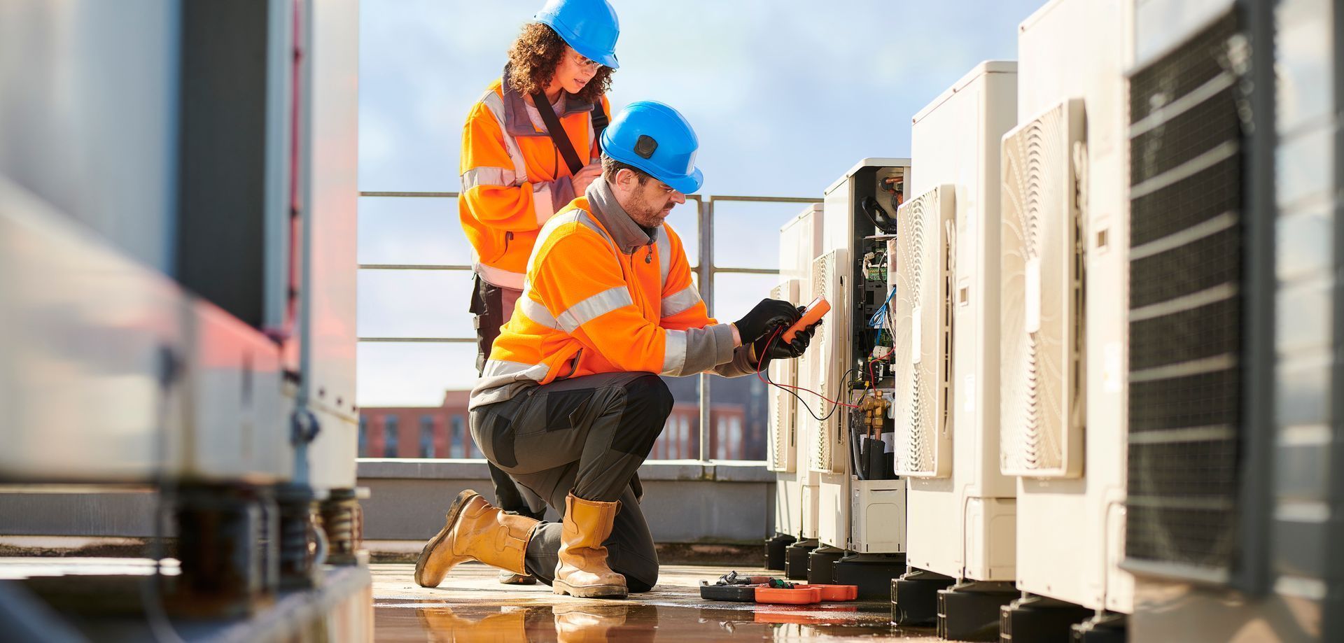 Two utility workers in orange safety gear inspecting rooftop electrical equipment at sunset