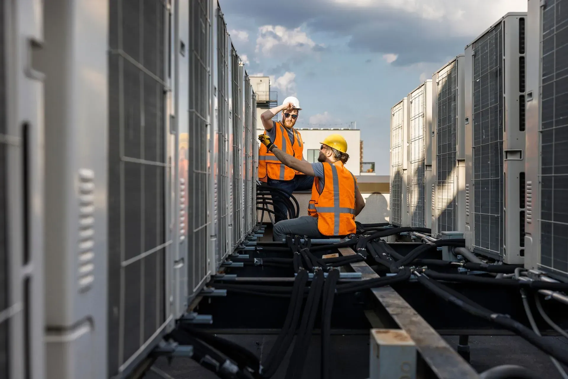 Workers in orange safety gear inspecting rooftop HVAC units and cables.