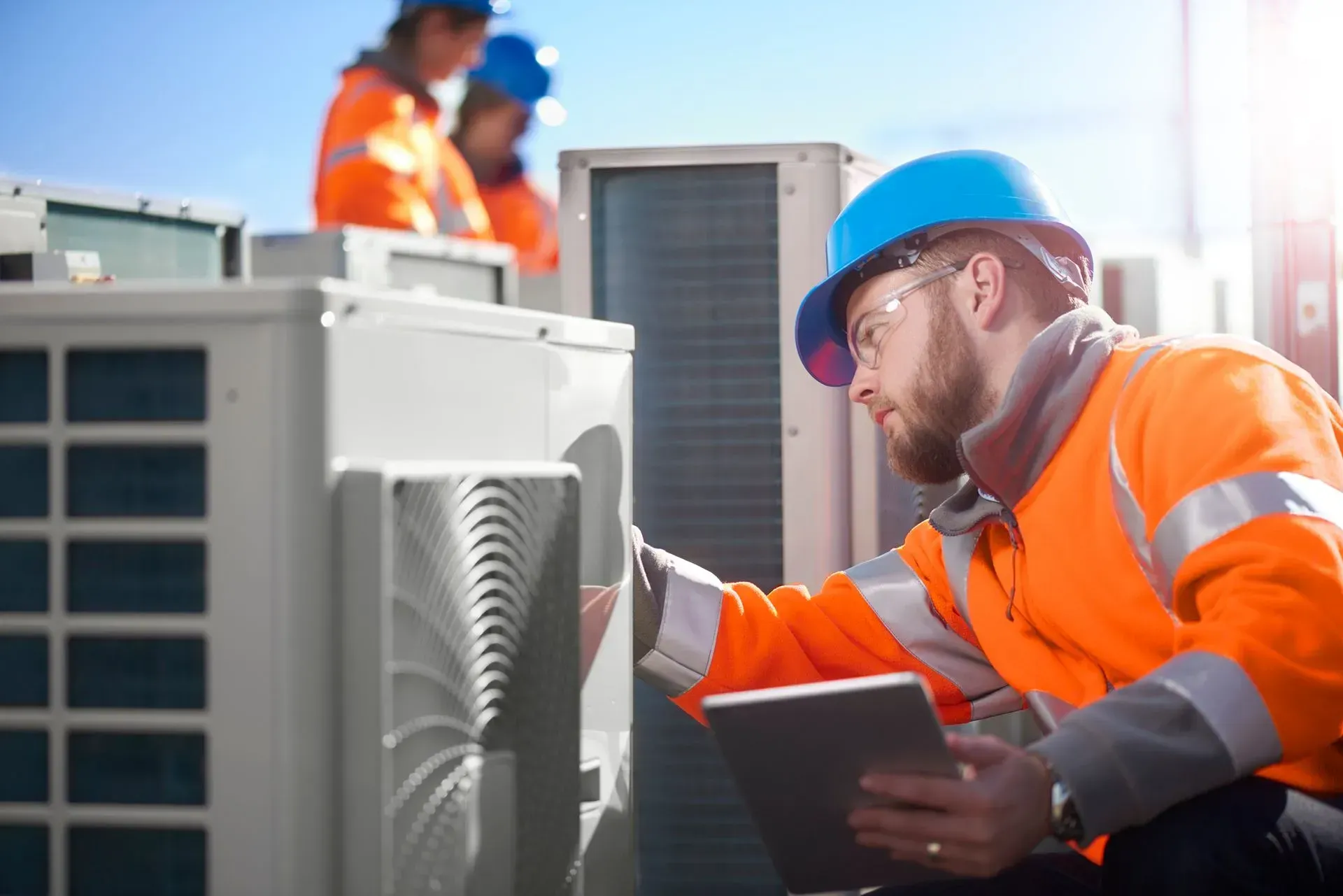 Technician in orange safety gear inspecting HVAC units outdoors, holding a tablet.