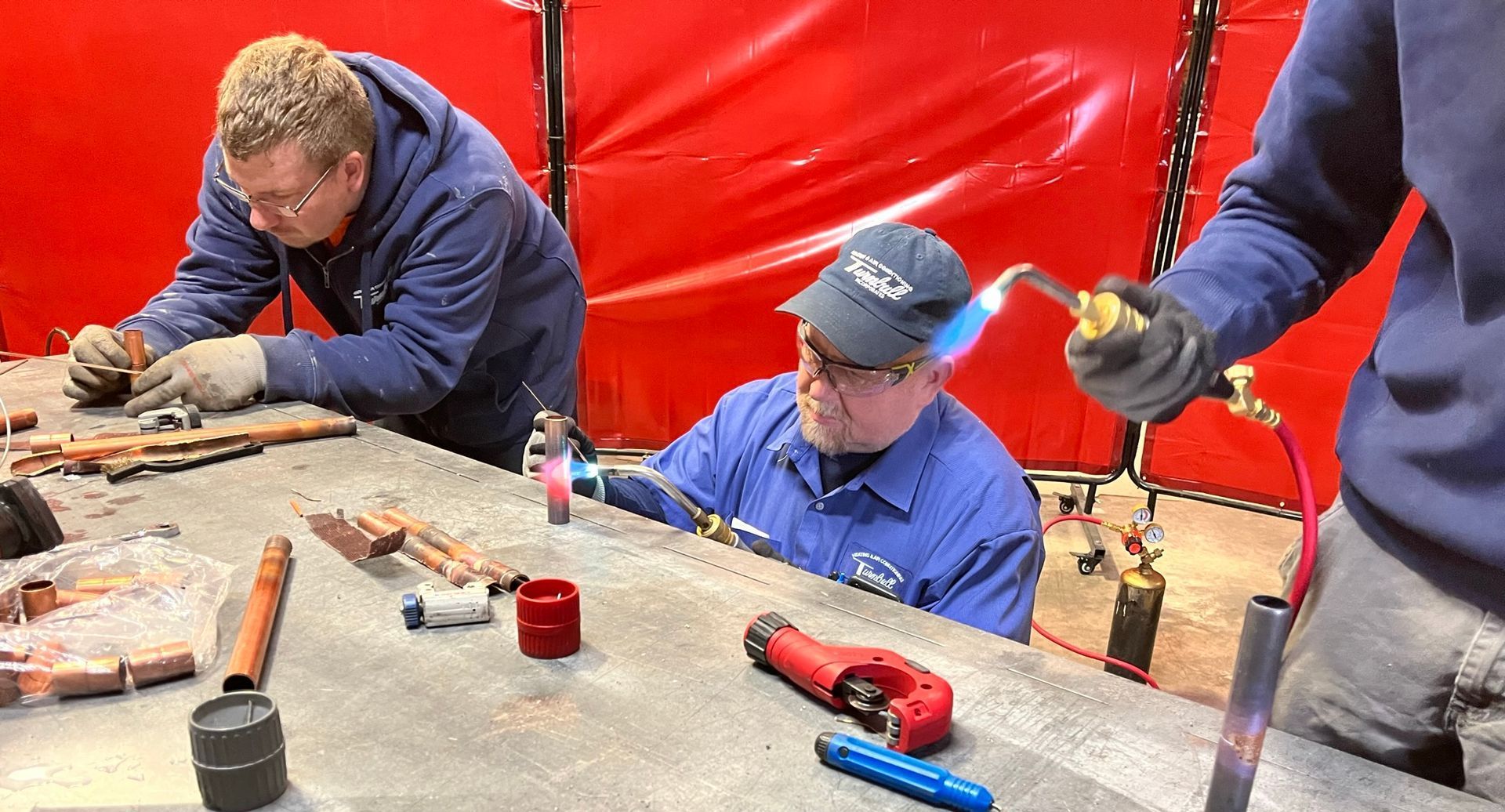 Three people soldering copper pipes in a workshop. One uses a torch, others work with tools. Red backdrop.