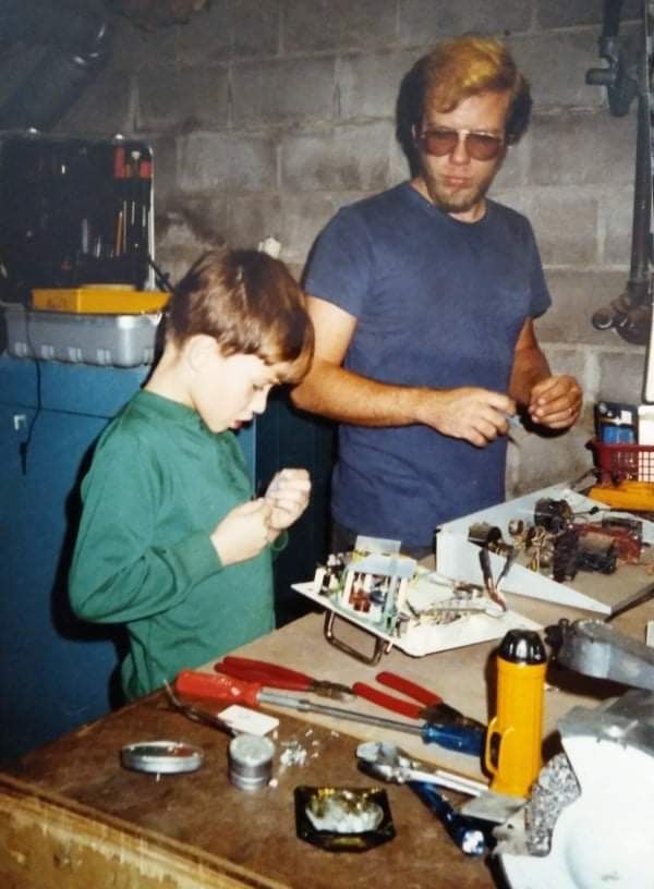 Boy and man working on electronics at a workbench in a basement, surrounded by tools and equipment.