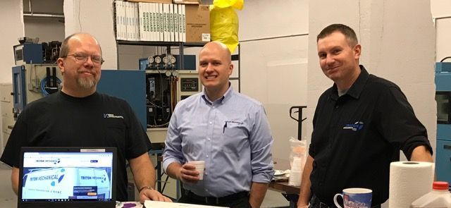 Three men smiling in a workshop with a laptop. One is holding a cup.