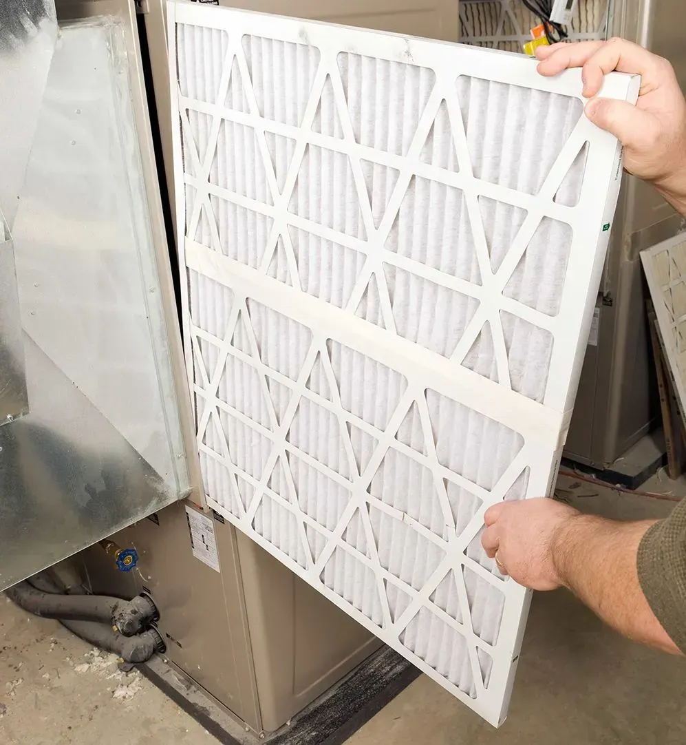 Hands removing a white HVAC air filter from a metal unit opening.