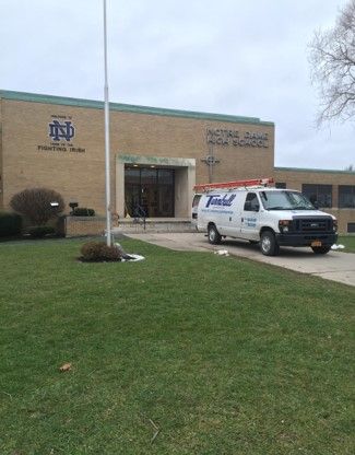 Exterior of a brick high school with a white van parked in front. Grass lawn, cloudy sky.