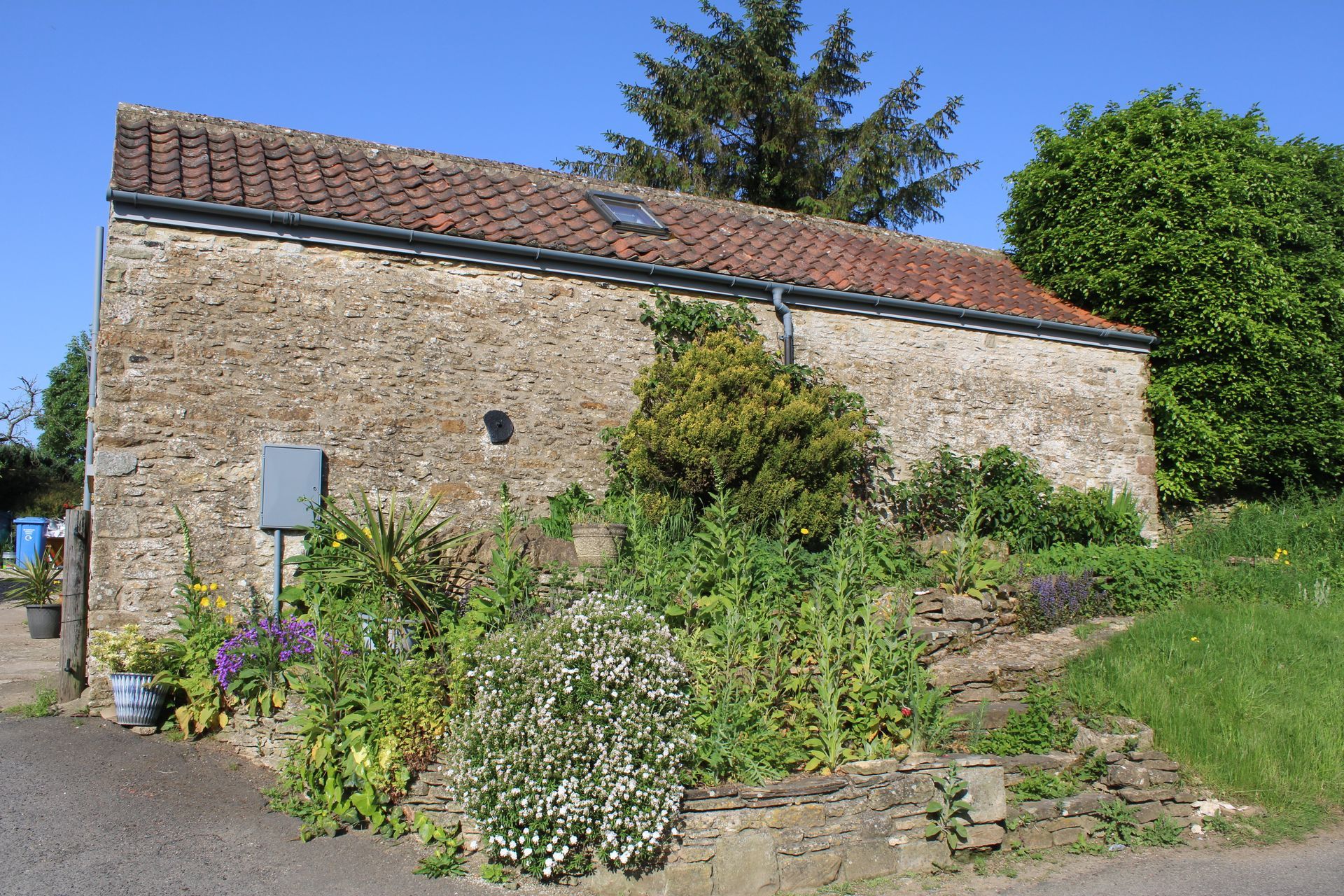 Small stone cottage with a tiled roof, surrounded by wildflowers and greenery in a sunny rural setting.
