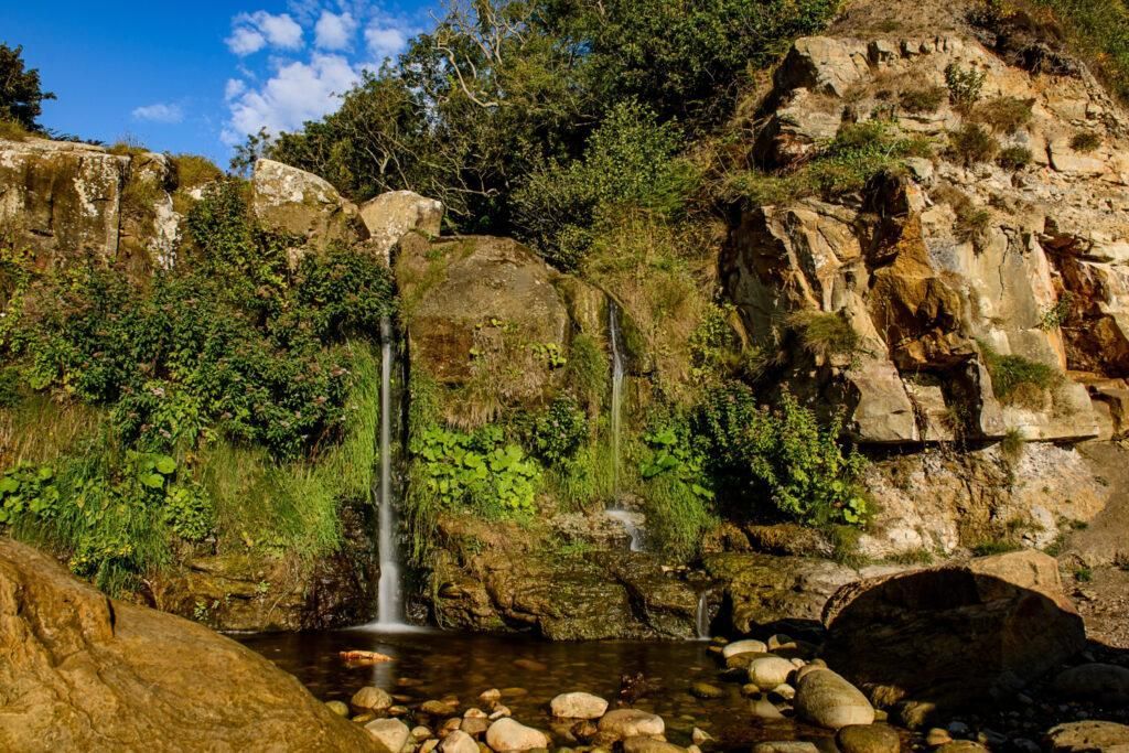 Small waterfall flowing into a rocky pool beside green moss-covered cliffs under a blue sky