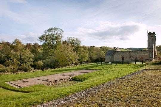 Grassy field with low stone ruins and a tall tower beside a tree-lined path under a cloudy sky