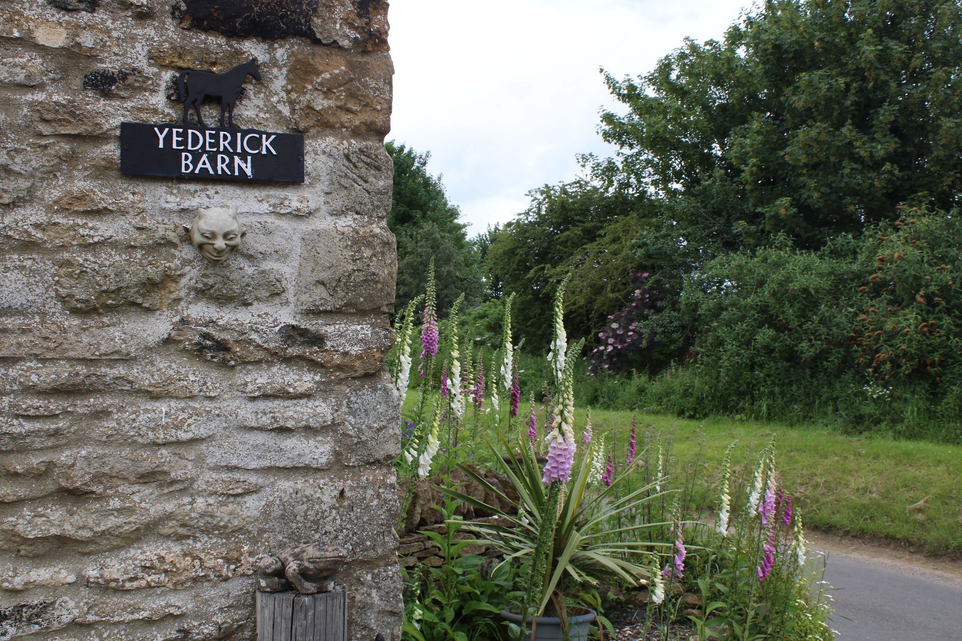 Stone barn wall with “YDELICK BARN” sign beside pink wildflowers and a country road