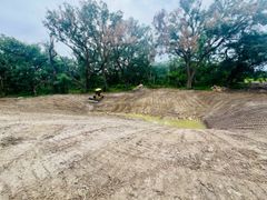 Graded dirt field beside trees and greenery under an overcast sky