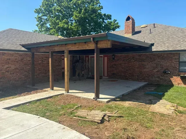 Brick house patio with a wooden awning, concrete slab, and small grassy yard