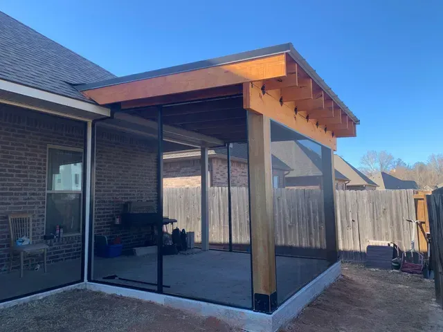 Covered patio with black mesh screens and wooden roof attached to a brick house in a backyard