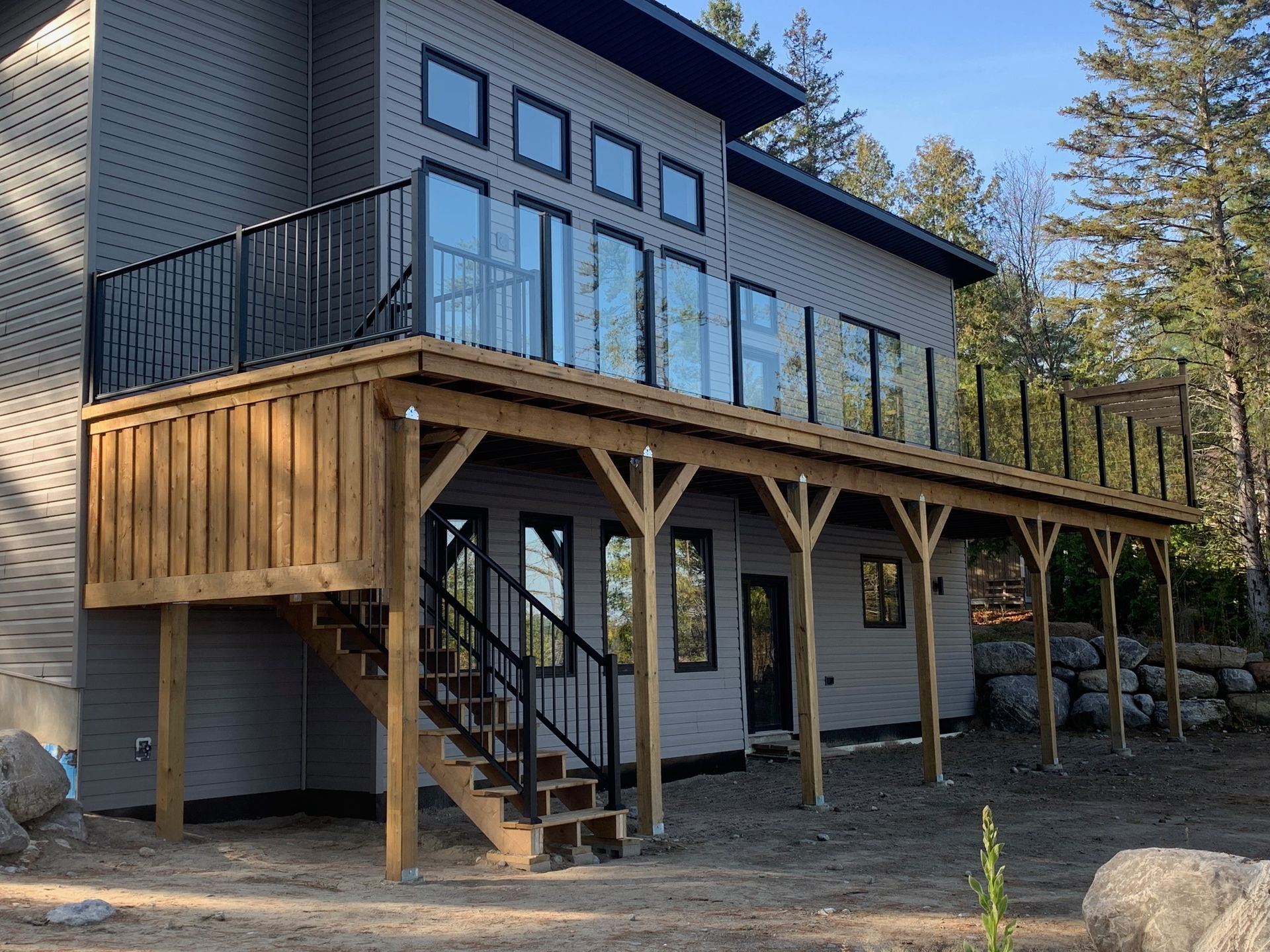 Modern gray house with a wooden deck, black railings, and stairs in a wooded area