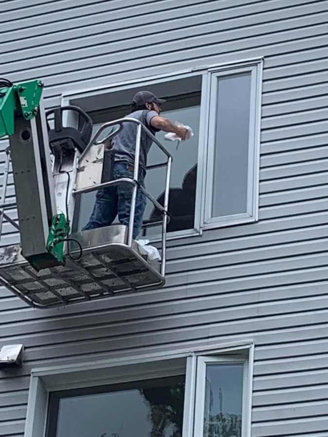 Worker in a lift painting or repairing an upstairs window on a gray-sided building.