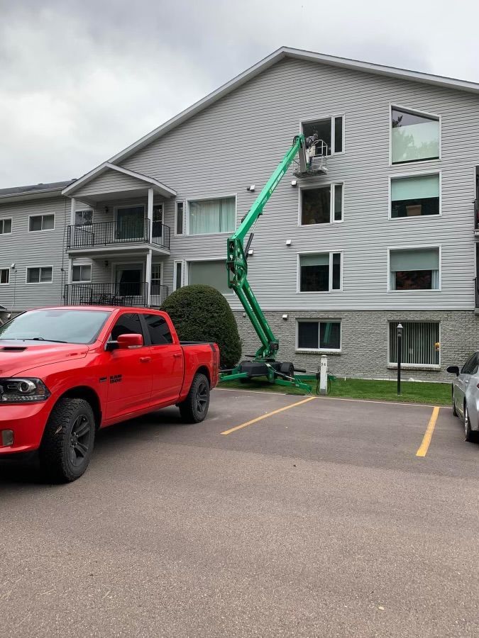 Green lift beside a gray apartment building, reaching to a second-floor window; red pickup parked in front.