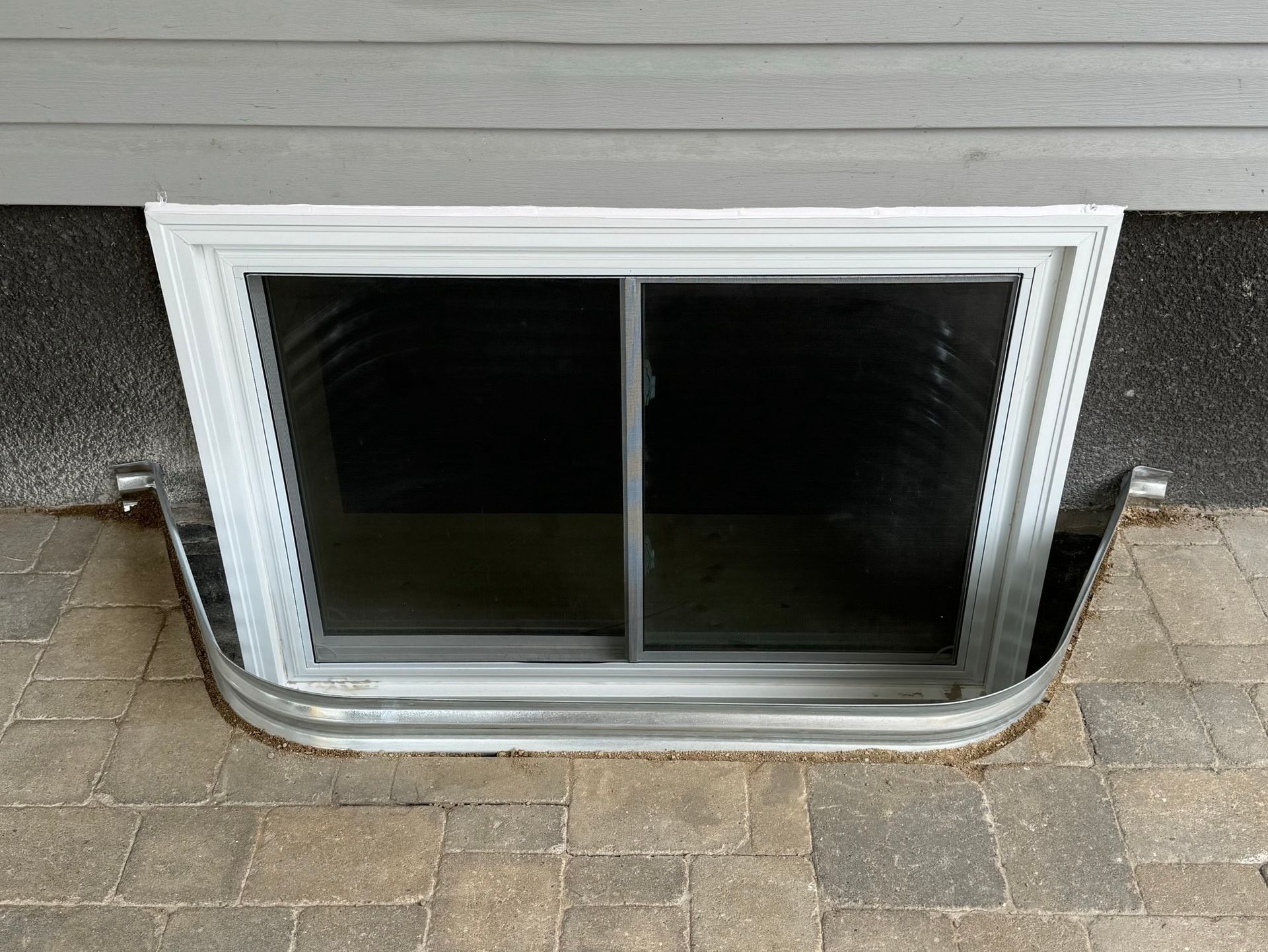 Small basement window with white trim set into a brick wall above stone pavers.