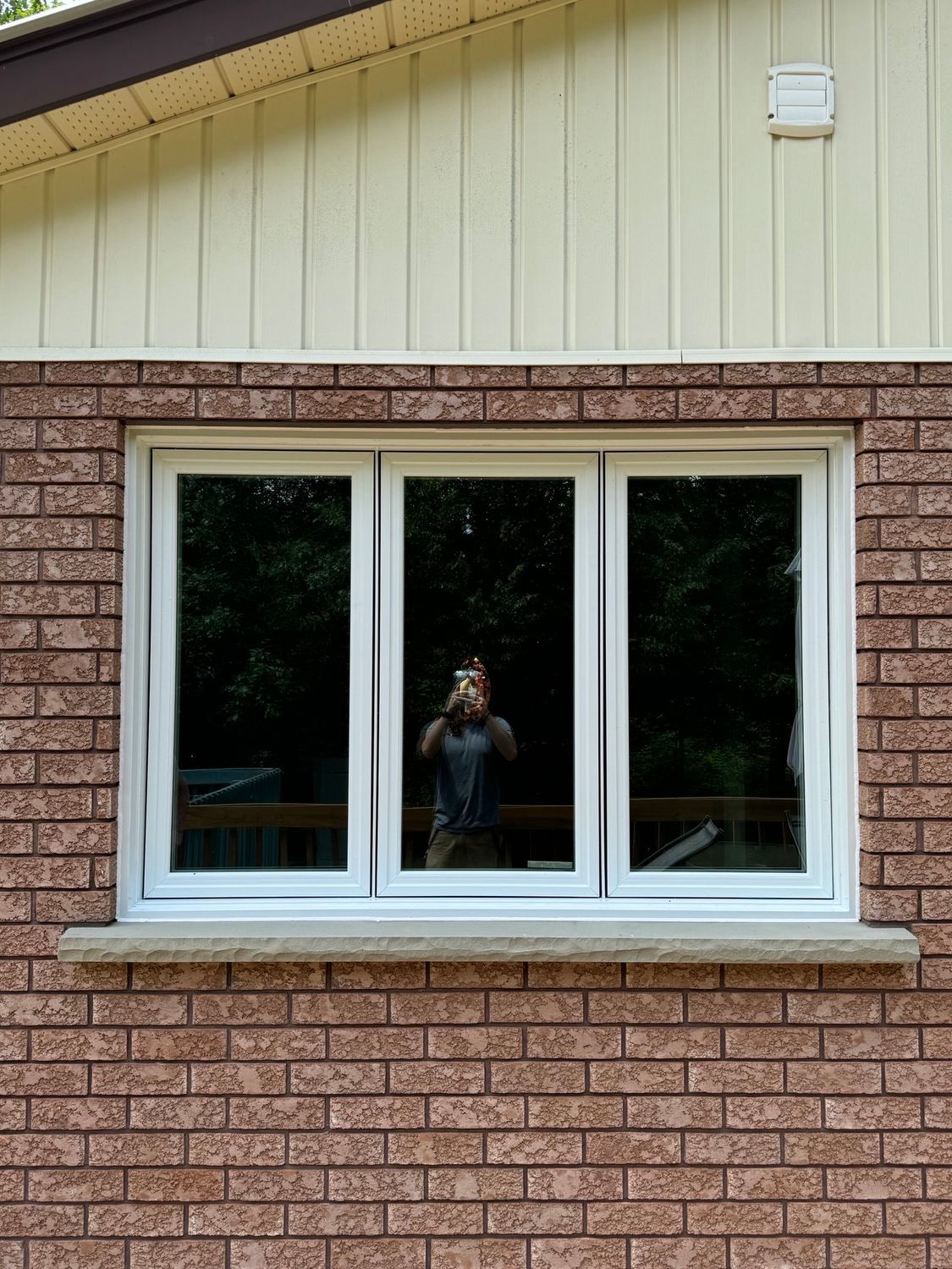 Brick house exterior with a white-framed window reflecting a person taking the photo.