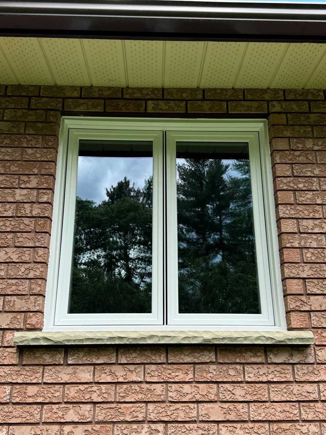 White-framed double window in a brick wall beneath a beige roof overhang, reflecting trees.