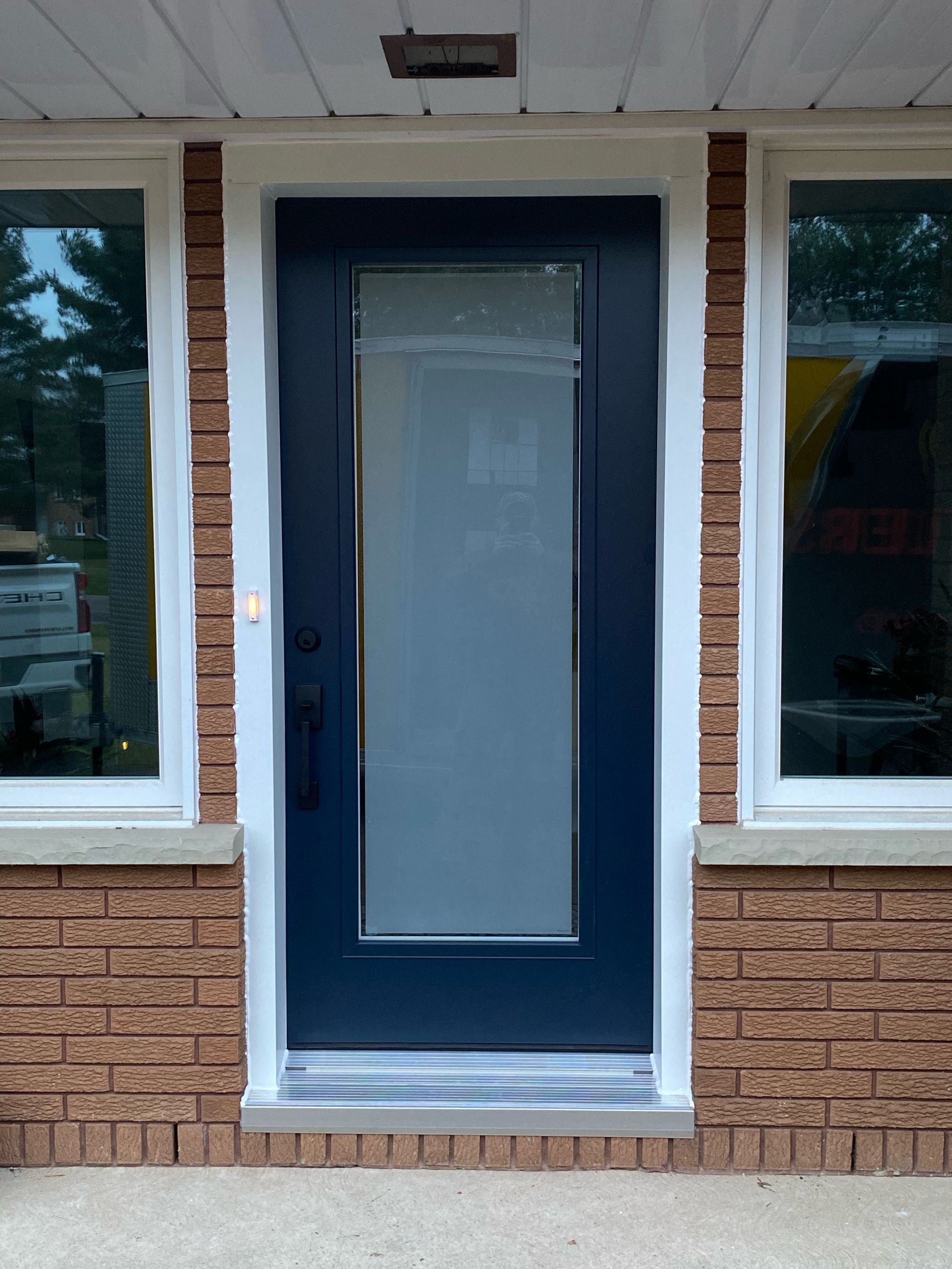 Blue front door with white glass panel between two windows on a brick house.
