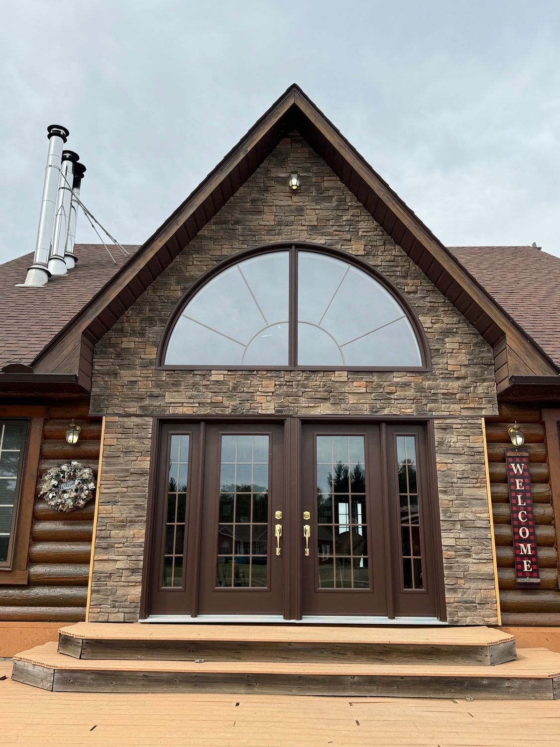 Stone-front house entrance with arched window, brown double doors, and log siding on the left
