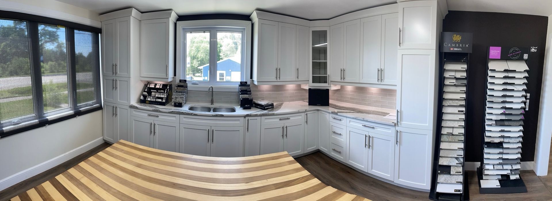 Bright kitchen with white cabinets, large windows, gray counters, and striped wood flooring.