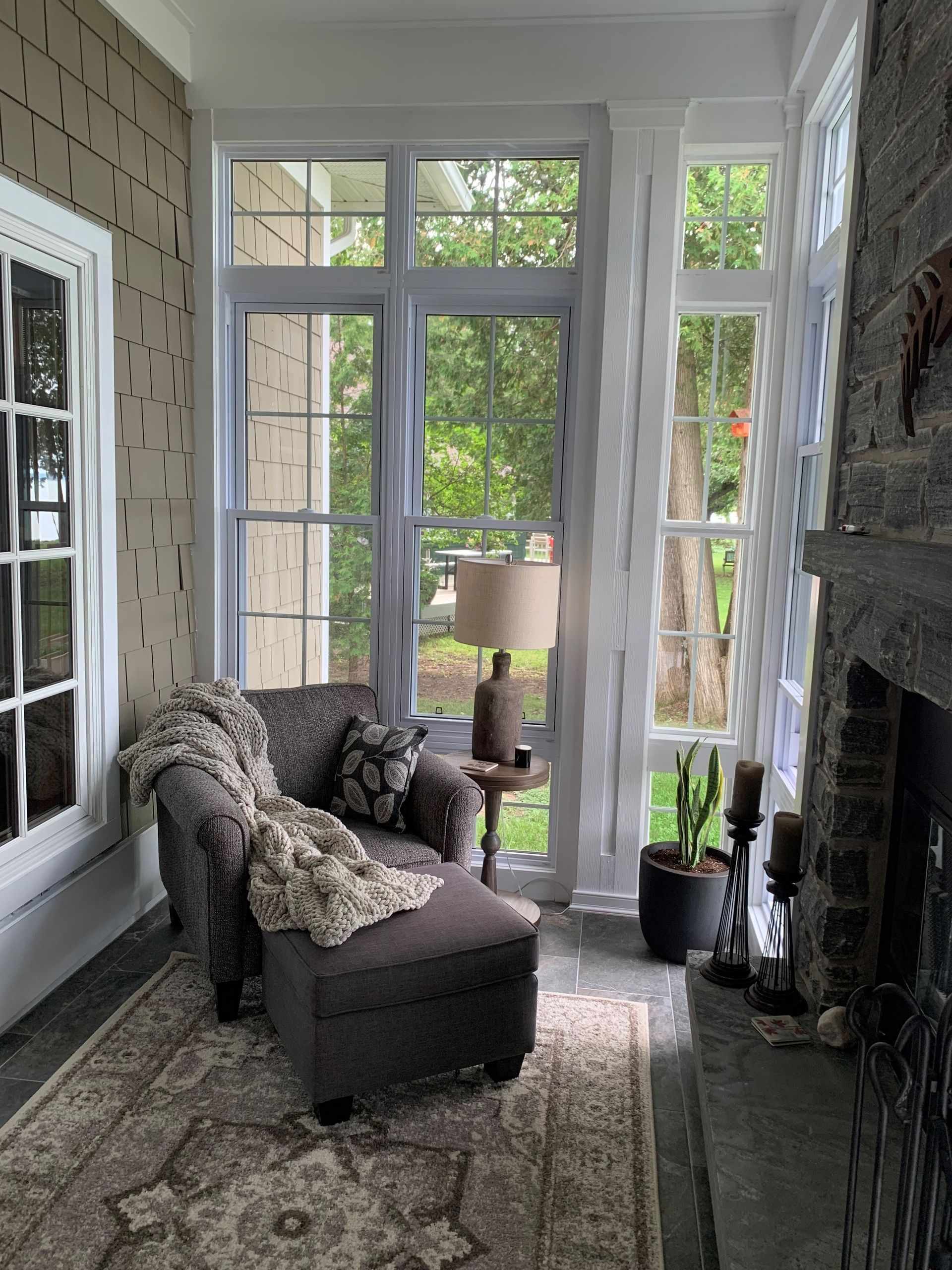 Bright sunroom with wicker chair, lamp, potted plant, and large windows overlooking greenery.