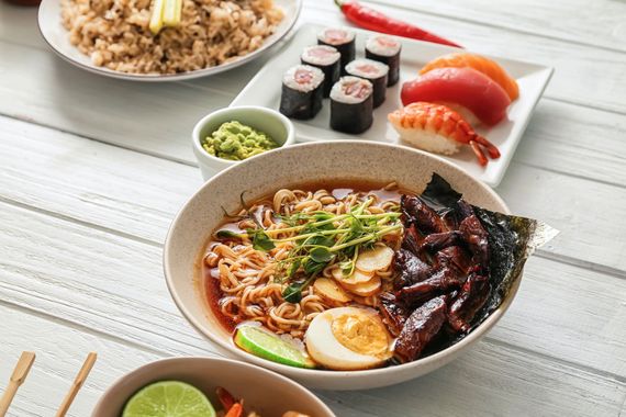 Japanese meal spread with ramen in foreground and sushi, rice, and condiments on a white table