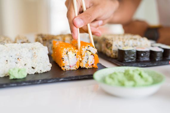 Hand using chopsticks to pick orange sushi rolls on a tray with wasabi and other rolls