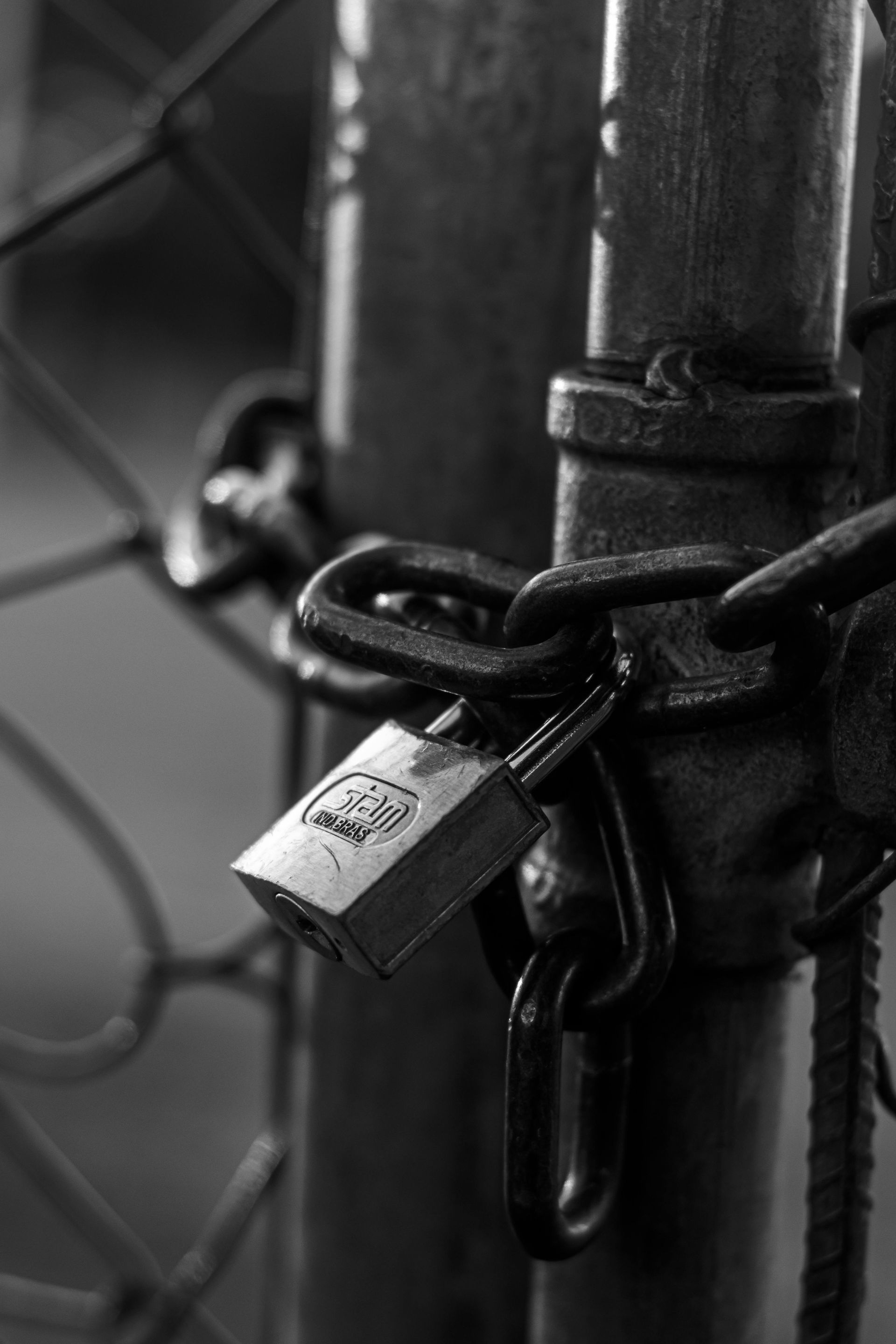 A black and white photo of a padlock on a chain link fence.