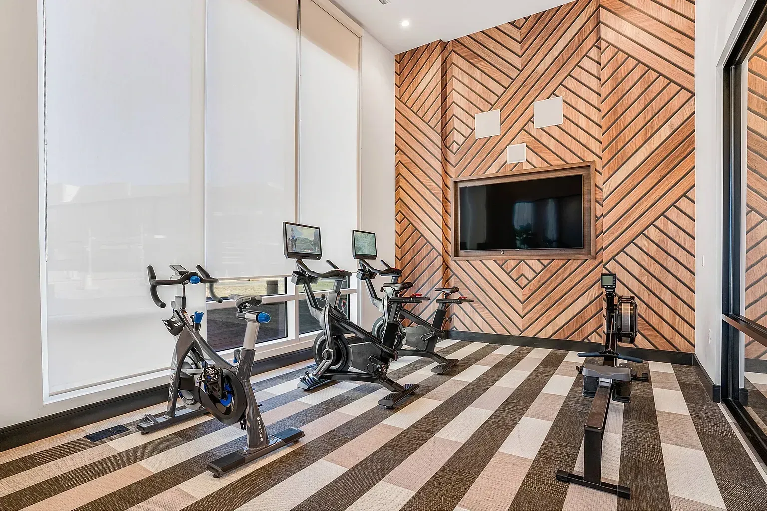 Spin bikes and rowing machine in a modern gym with wood accent wall and TV.