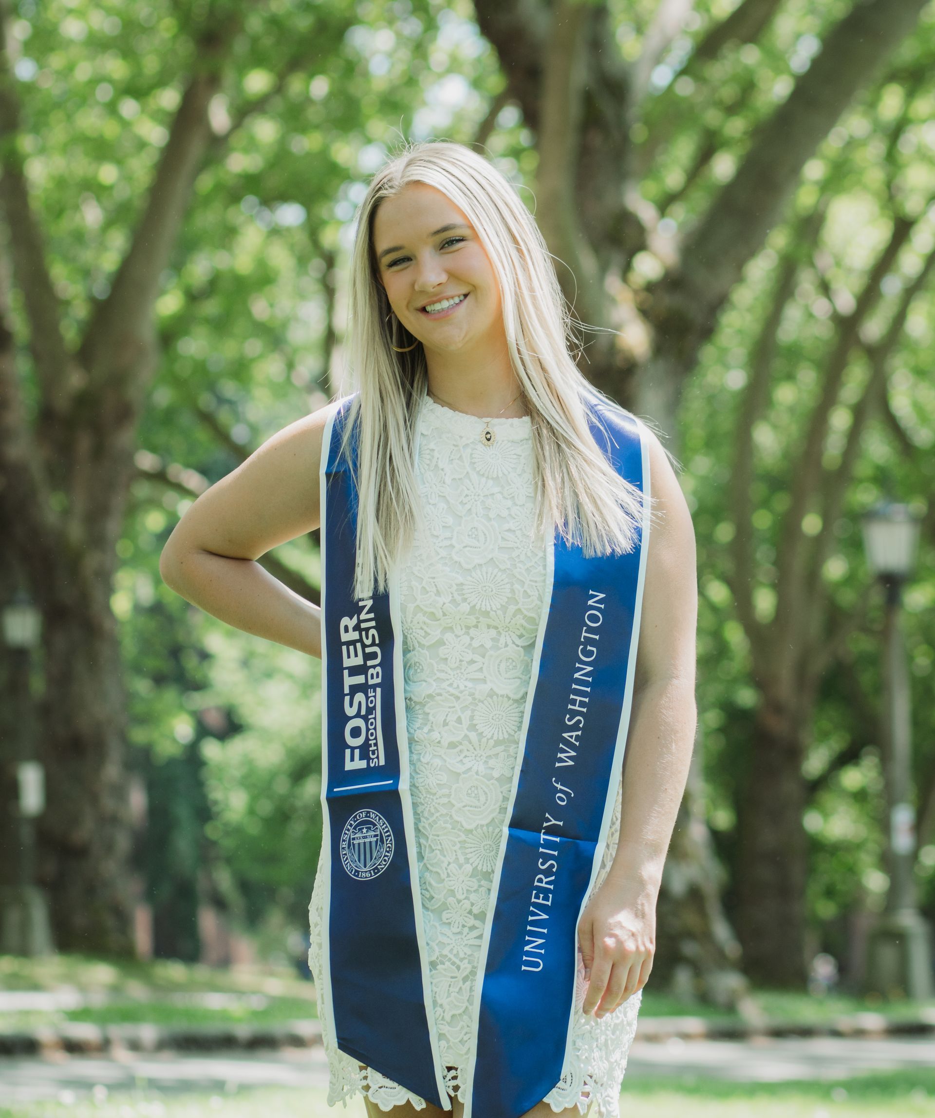A woman in a white dress and blue sash is standing in a park.