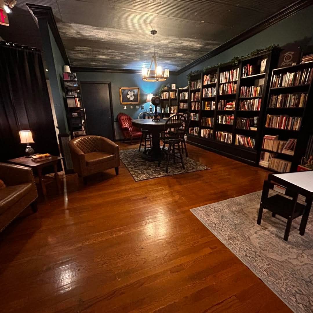 A dimly lit library or study with wooden floors, dark bookshelves, leather chairs, and a round table under a chandelier.