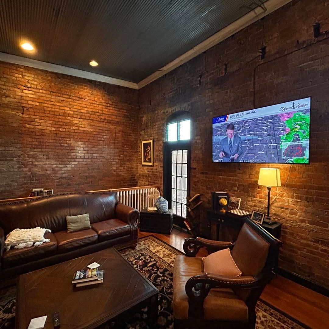A room with exposed brick walls, a leather sofa, an armchair, a coffee table, and a wall-mounted TV showing a weather map.