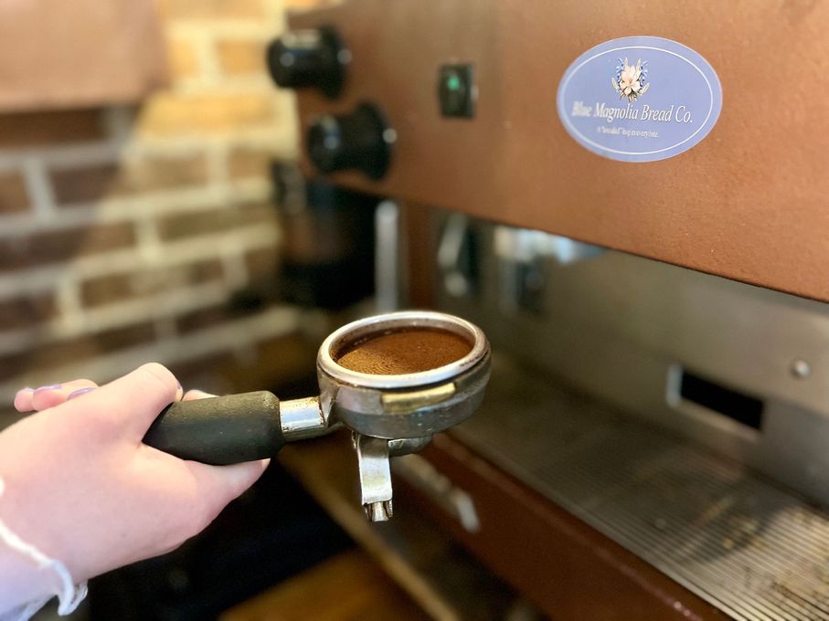 A person holds a portafilter filled with ground coffee in front of an espresso machine with a light-blue circular logo.