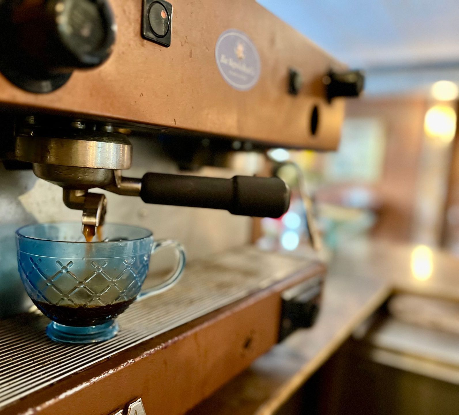 Espresso drips from a professional machine into a textured blue glass cup on a metal counter.