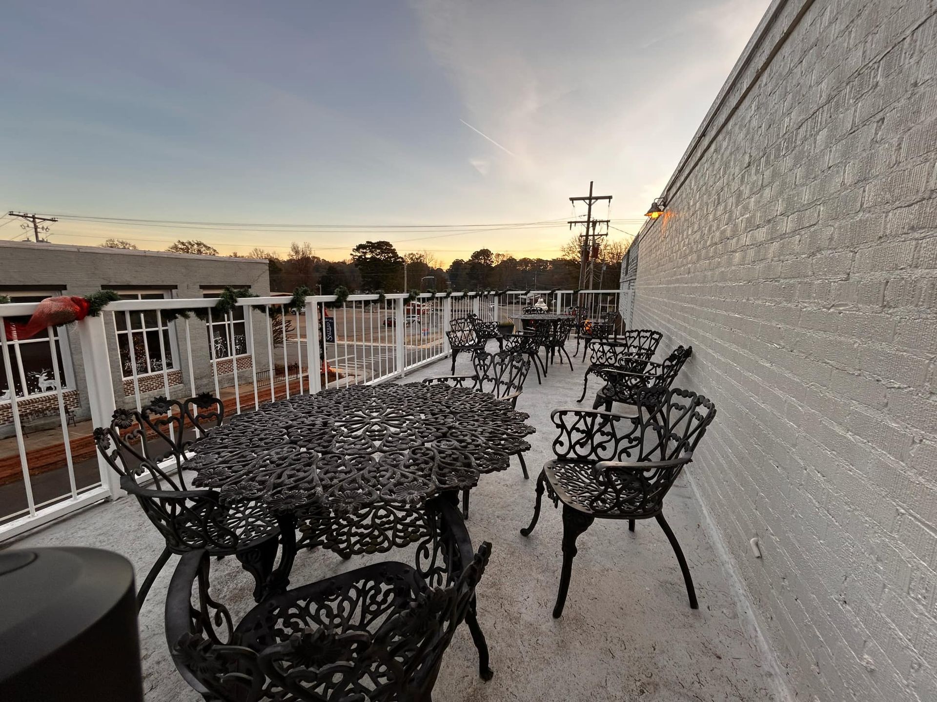 Outdoor patio featuring wrought iron tables and chairs alongside a long, white brick wall at sunset.
