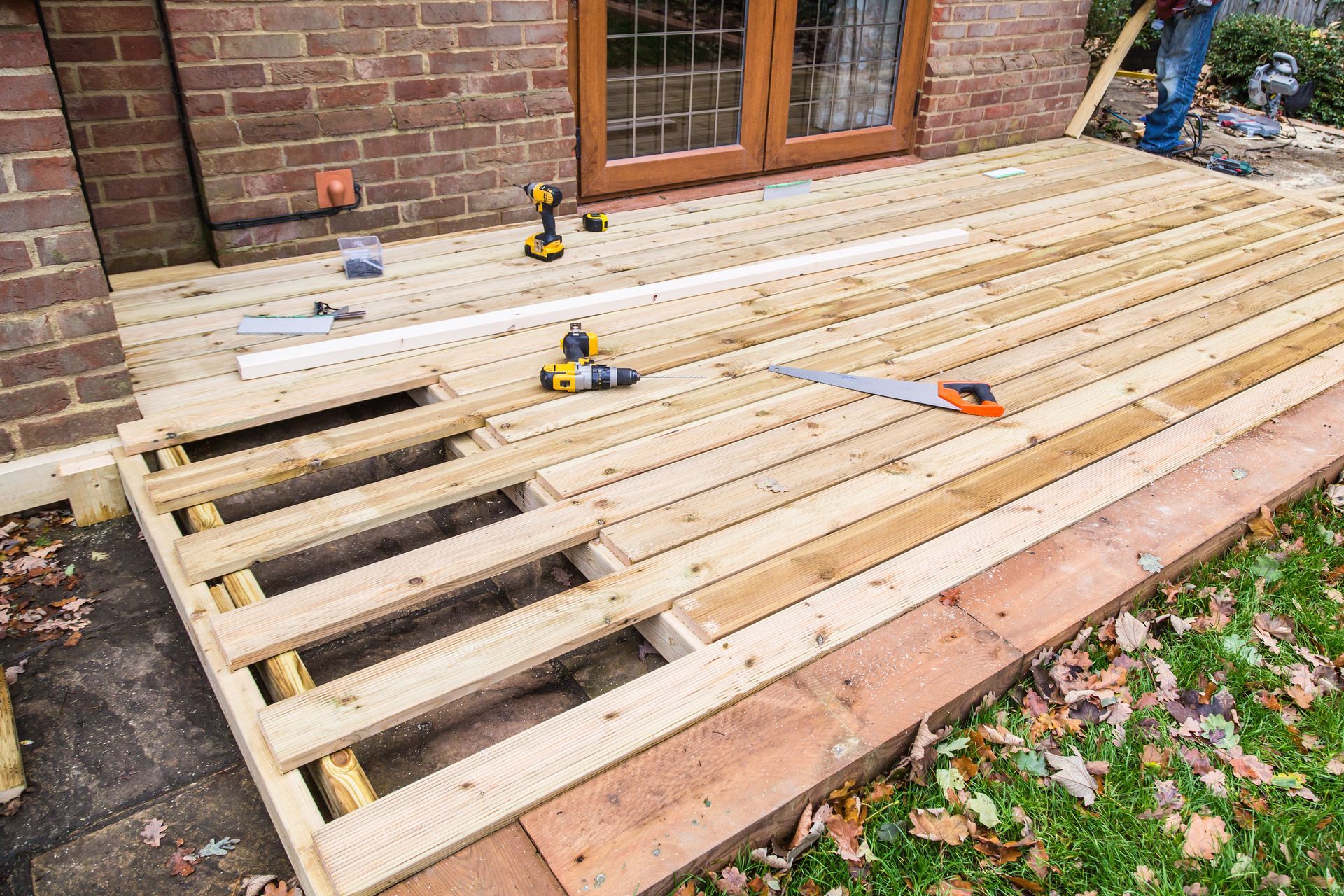 Partially built wooden deck beside a brick house, with tools and open joists visible Partially built wooden deck beside a brick house, with tools and open joists visible