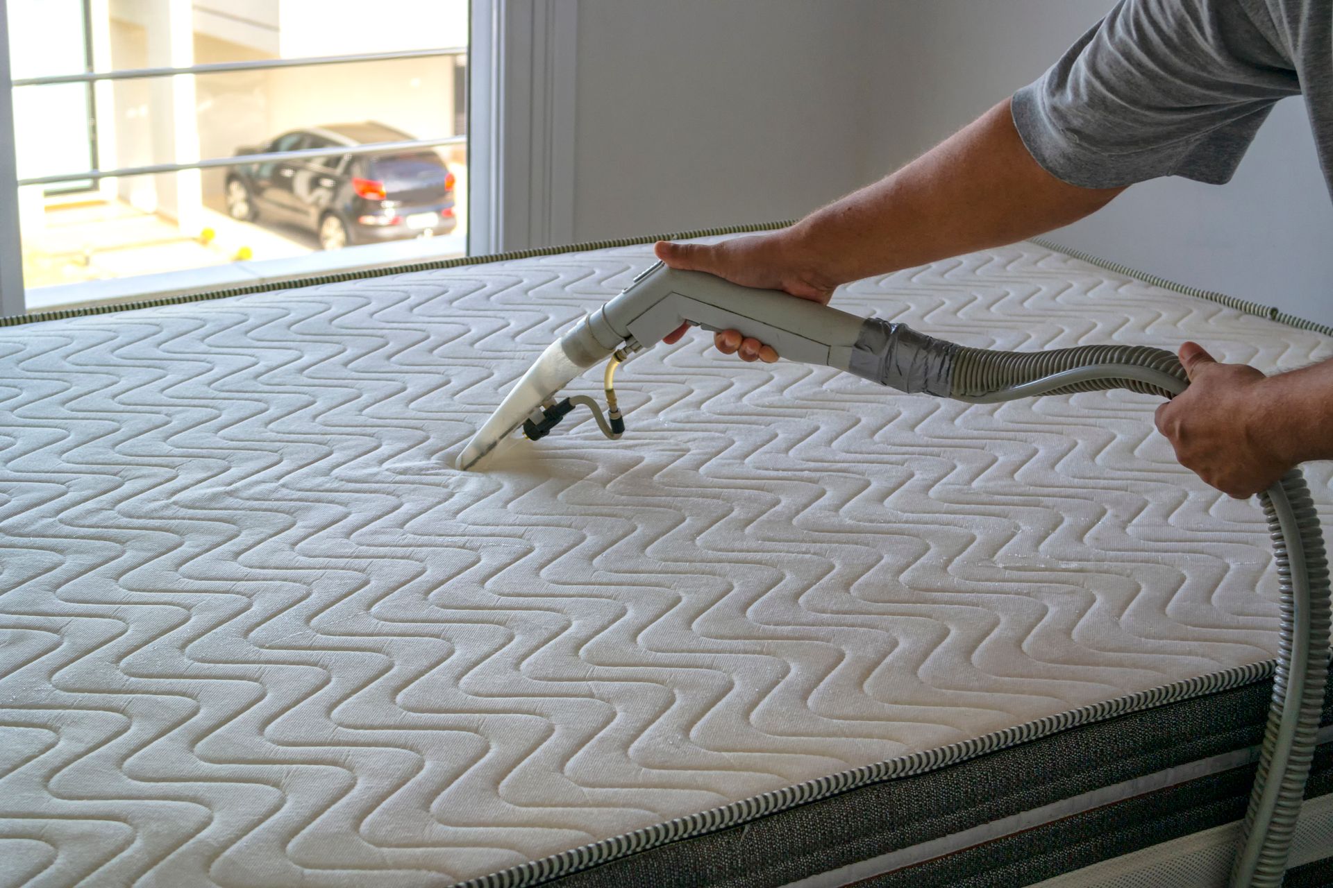 Person vacuuming a mattress; white and grey colors; near a window; cleaning action.